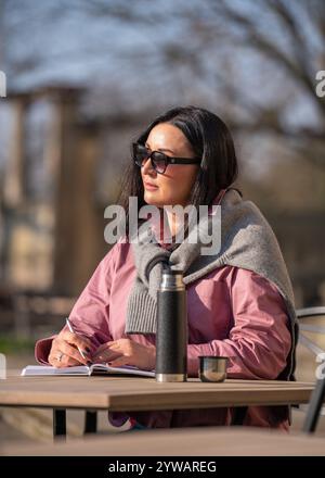 Hübsche Frau, die in einem Café im Freien sitzt und Notizen in Notizbuch macht. In der Nähe auf dem Tisch befindet sich eine Thermoskanne mit brennendem Getränk. Brünette arbeitet ferngesteuert, denk Stockfoto