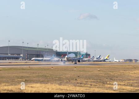 Ein Verkehrsflugzeug landet auf der Landebahn mit sichtbarem Rauch aus seinen Reifen, umgeben von anderen Flugzeugen und dem Flughafenterminal unter einem klaren Blau. Stockfoto