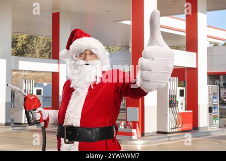 Der Weihnachtsmann hält eine Kraftstoffspritzpistole und drückt an einer Tankstelle die Daumen hoch Stockfoto