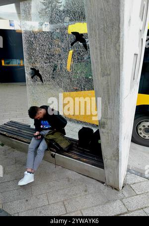 Junger Mann, der auf das Smartphone blickte, während er vor einer großen Scheibe aus beschädigtem Glas im Busbahnhof lenti zala, ungarn, saß Stockfoto