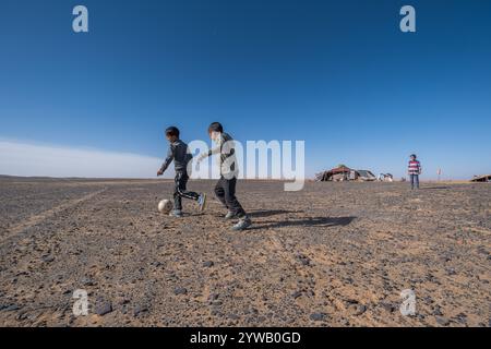 Drei Kinder spielen Fußball in der Weite der felsigen marokkanischen Wüste nahe Merzouga. Im Hintergrund befinden sich die typischen Nomadenzelte im ca. Stockfoto