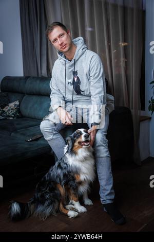 Ein junger Mann in einem hellgrauen Hoodie sitzt auf einem Sofaarm, mit einem australischen Schäferhund auf dem Boden. Der Raum verfügt über sanfte Beleuchtung und transparente Vorhänge. Stockfoto