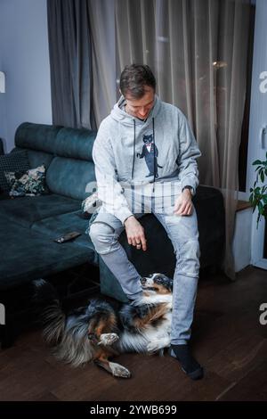 Ein junger Mann in einem grauen Hoodie mit einer Katzengrafik sitzt auf einem dunkelgrünen Sofa und interagiert spielerisch mit einem dreifarbigen Hund auf einem Holzboden in einem sanft beleuchteten Ro Stockfoto