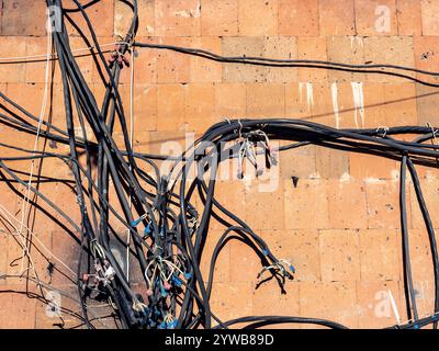 Unordentliche elektrische Kabel und Drähte an der Außenwand des Gebäudes, bedeckt mit orangen Fliesen aus armenischem Tuff in Jerewan Stadt an sonnigen Tagen Stockfoto