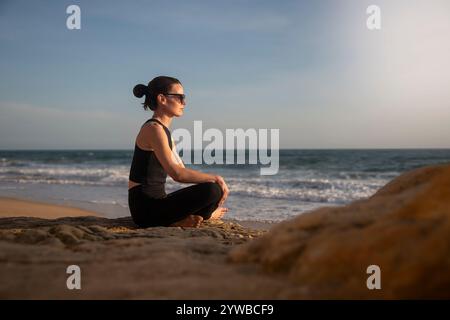 Sportliche Frau, die mit überkreuzten Beinen am Strand sitzt und den Sonnenuntergang beobachtet. Von allem wegkommen. Stockfoto