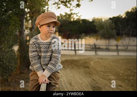 Junge in einem alten Hut und Pullover auf dem Zaun einer Reitarena Stockfoto