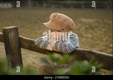 Junge in einem alten Hut und Pullover auf dem Zaun einer Reitarena Stockfoto