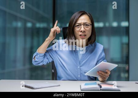 Eine asiatische Geschäftsfrau, die starke Emotionen zeigt, während sie in einem modernen Büro ein Tablet hält. Ihre selbstbewusste Geste deutet auf eine ernsthafte Diskussion oder eine wichtige Entscheidungssituation hin. Stockfoto