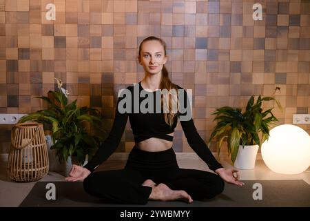 Ein athletisches Mädchen in schwarzer Sportkleidung sitzt in Lotusposition bei einem Yoga-Kurs in einem Fitnessraum inmitten grüner Hallenpflanzen. Stockfoto