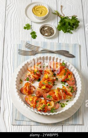 Uunimakkara, gebacken im Ofen Würstchen mit Käse, Tomaten, Koriander, Senf in Auflaufform auf weißem Holztisch mit Zutaten, finnischer Rückgabe Stockfoto