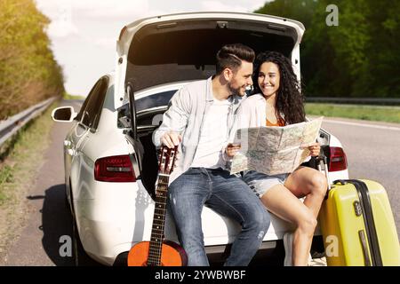 Junges Paar, das mit dem Auto unterwegs ist, im Kofferraum mit Koffer und Gitarre sitzt, Karte hält, Ziel überprüft Stockfoto