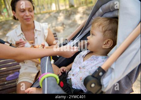 Mutter und Kind verbringen einen süßen Moment, während das Baby im Park ein Eis genießt. Sie sitzen auf einer Bank in einem entspannten, sonnigen Outdoor-Set Stockfoto