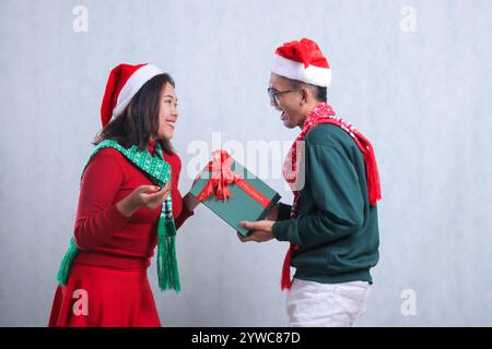 Zwei junge asiatische Männer, die weihnachtspullover, weihnachtsmützen und Tücher tragen, sich glücklich anschauen, sein Freund gab ihm ein Green Box Geschenk, isolieren Stockfoto
