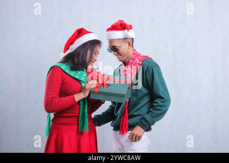 Porträt eines jungen indonesischen Paares, das Weihnachtspullover, Weihnachtsmützen und Tücher trägt, fröhlich auf die Geschenkbox blickt, isoliert auf weißem Hintergrund Stockfoto
