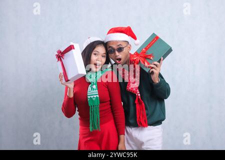 Geste eines jungen Paares mit Weihnachtspullover, Weihnachtsmütze und Schal, überrascht auf der Vorderseite mit roter Schachtel und weißer Schachtel mit Weihnachtsgeschenken, iso Stockfoto