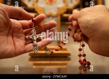 Ältere kaukasische Frau mit faltigen Händen, die einen roten Rosenkranz in der rechten Hand und das Kruzifix in der linken Hand hält. Fotografiert mit einem flachen Stockfoto