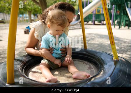 Eine fröhliche Mutter hält ihr Kind sanft fest, während sie auf einer Reifenschaukel in einem lebhaften Park spielt. Stockfoto