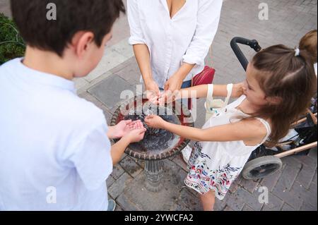 Eine Familie versammelt sich an einem öffentlichen Brunnen, planscht und genießt das kühle Wasser an einem sonnigen Tag. Kinder und Erwachsene genießen Spaß und Erfrischungen Stockfoto