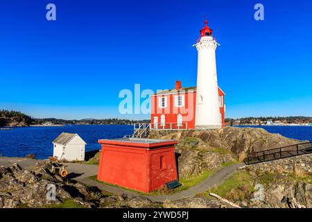 Ein roter Leuchtturm steht an einer felsigen Küste. Der Leuchtturm ist von einem kleinen Gebäude und einem Pfad umgeben, der zu ihm führt. Der Himmel ist klar und blau, und Stockfoto