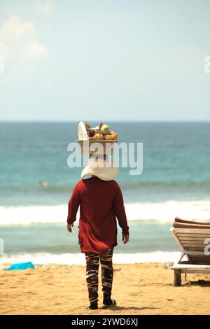 Eine Frau trägt frisches Obst auf dem Kopf, während sie am Strand von Kuta in Kuta, Badung, Bali, Indonesien, Käufer findet. Stockfoto