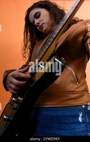 Junge Musikerin mit lockigen Haaren und Lederjacke, die elektrische Gitarre auf orangem Hintergrund spielt Stockfoto