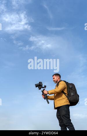 Ein professioneller Videofilmer, der einen kardanalen Kamerabildstabilisator verwendet, nimmt draußen unter blauem Himmel auf Stockfoto