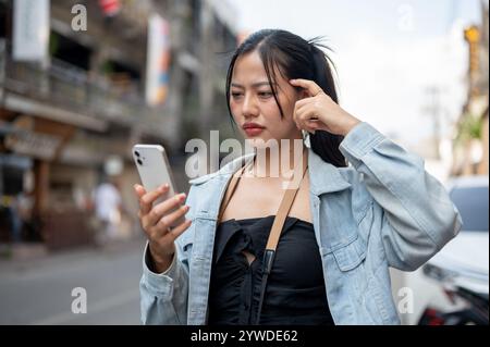 Eine verwirrte asiatische Touristin in einer Jeansjacke, die ihr Smartphone benutzt, um die Richtung auf der Karte-App zu überprüfen, während sie die Straße entlang läuft und sich nicht mehr anfühlt Stockfoto