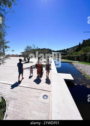 Eine Familie genießt einen gemütlichen Spaziergang entlang eines sonnendurchfluteten Pfades neben einem ruhigen Teich, umgeben von wunderschönem Grün. Die Szene fängt eine friedliche und fröhliche Atmosphäre ein Stockfoto