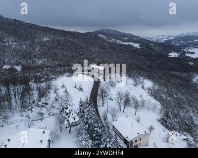 Schneebedeckte Straße schlängelt sich durch eine ruhige Berglandschaft, die zu Häusern führt, die zwischen schneebedeckten Bäumen liegen und eine ruhige Winterszene bieten. Stockfoto