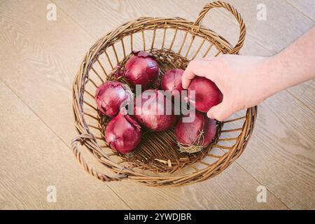 Eine Hand reicht in einen braunen Korb mit roten Zwiebeln, der auf einem Holzboden liegt. Stockfoto