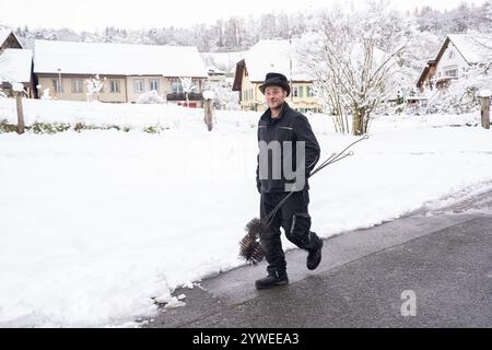 Ein fröhlicher Schornsteinfeger geht selbstbewusst durch eine schneebedeckte Dorfstraße und trägt seine Putzwerkzeuge. Die heitere Winterszene zeigt ein Q Stockfoto