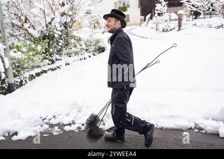 Ein Schornsteinfeger geht durch ein schneebedecktes Viertel, mit Werkzeugen. Das Bild zeigt einen Winterarbeitstag mit traditionellem Schornsteinfeger Stockfoto