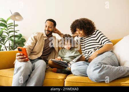 Familie sitzt entspannt auf einer gelben Couch und verbringt Zeit zu Hause. Mutter und Sohn lesen und der Vater benutzt ein Smartphone. Stockfoto