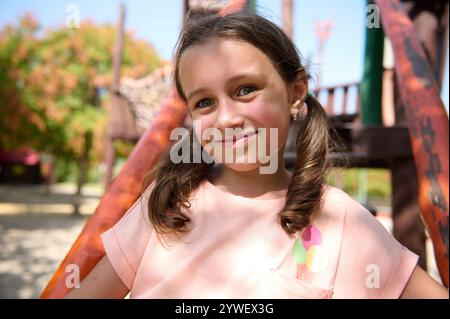 Ein fröhliches junges Mädchen mit Zöpfen lächelt hell, während es auf einem sonnigen Spielplatz spielt. Stockfoto