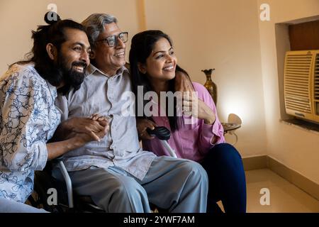 Glückliches, junges indisches Paar, das zu Hause mit einem behinderten älteren Vater auf dem Rollstuhl sitzt. Elterntag- oder Vatertag-Konzept. Stockfoto