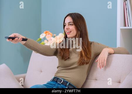 Frau, die auf dem Sofa sitzt, die Fernbedienung hält und drinnen lächelt Stockfoto
