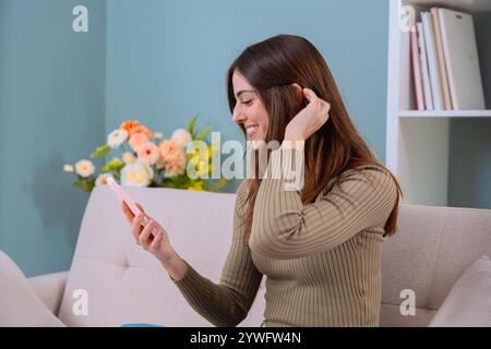 Lächelnde Frau, die ein Smartphone benutzt, während sie auf einem gemütlichen Sofa sitzt. Stockfoto