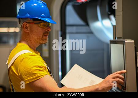 Industrieingenieur, der eine cnc-Fräsmaschine mit einem numerischen Bedienfeld verwaltet und eine technische Zeichnung hält. Stockfoto