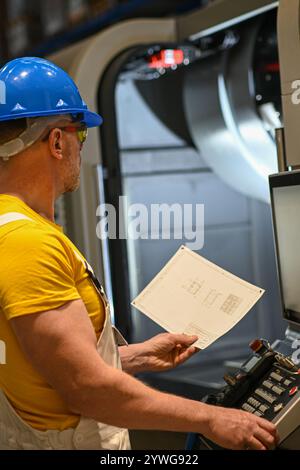 Industrieingenieur, der eine cnc-Fräsmaschine mit einem numerischen Bedienfeld verwaltet und eine technische Zeichnung hält. Stockfoto