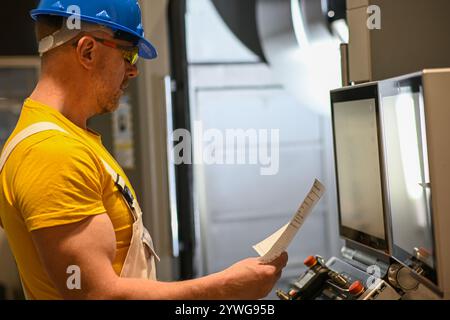 Industrieingenieur, der eine cnc-Fräsmaschine mit einem numerischen Bedienfeld verwaltet und eine technische Zeichnung hält. Stockfoto