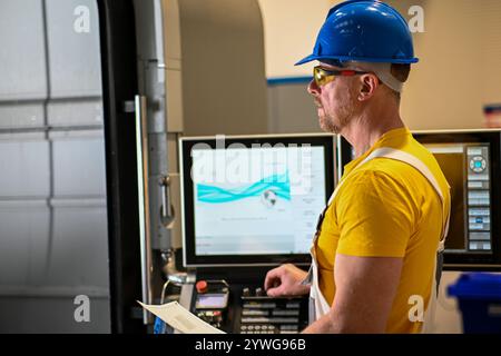 Industrieingenieur, der eine cnc-Fräsmaschine mit einem numerischen Bedienfeld verwaltet und eine technische Zeichnung hält. Stockfoto