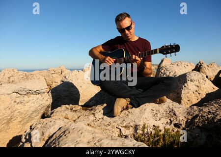 Ein Mann in einem dunklen T-Shirt spielt Akustikgitarre auf einem felsigen Gebiet am Meer Stockfoto