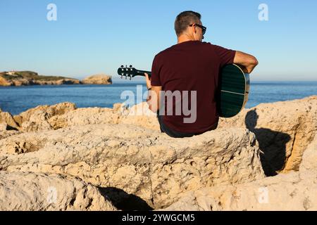 Ein Mann in einem dunklen T-Shirt spielt Akustikgitarre auf einem felsigen Gebiet am Meer Stockfoto