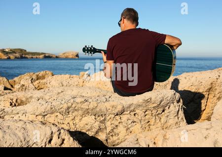 Ein Mann in einem dunklen T-Shirt spielt Akustikgitarre auf einem felsigen Gebiet am Meer Stockfoto