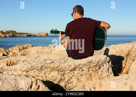 Ein Mann in einem dunklen T-Shirt spielt Akustikgitarre auf einem felsigen Gebiet am Meer Stockfoto