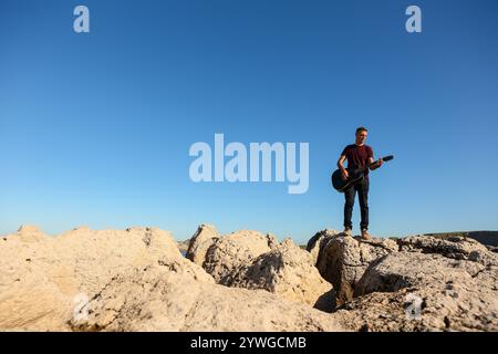 Ein Mann in einem dunklen T-Shirt spielt Akustikgitarre auf einem felsigen Gebiet am Meer Stockfoto