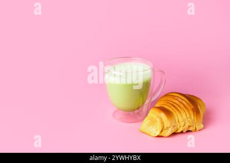 Matcha Latte mit einem Schaum in Glasbecher mit Croissant auf hellrosa Hintergrund mit Kopierraum Stockfoto