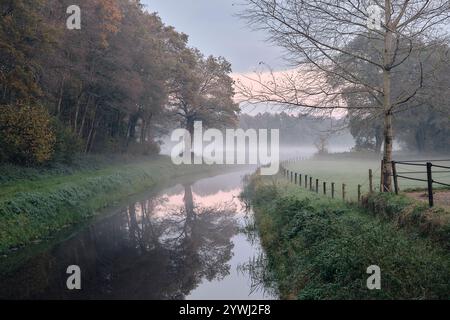 Ein malerischer Blick auf einen kleinen Kanal im ländlichen Drenthe, mit nebligen frühen Sonnenuntergang, Herbstbäumen und Wiesen in einer ruhigen natürlichen Umgebung. Stockfoto