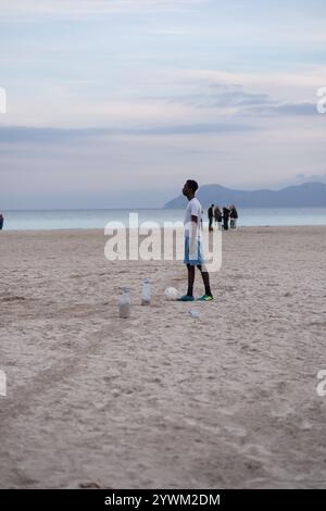 Junger Afroamerikaner mit einem Ball am Strand. Der Typ spielt Fußball am Sandstrand. Stockfoto