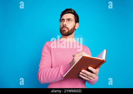 Selbstbewusster junger Mann in rosa Pullover, der Notizbuch hält und entschlossen vor blauem Hintergrund denkt Stockfoto